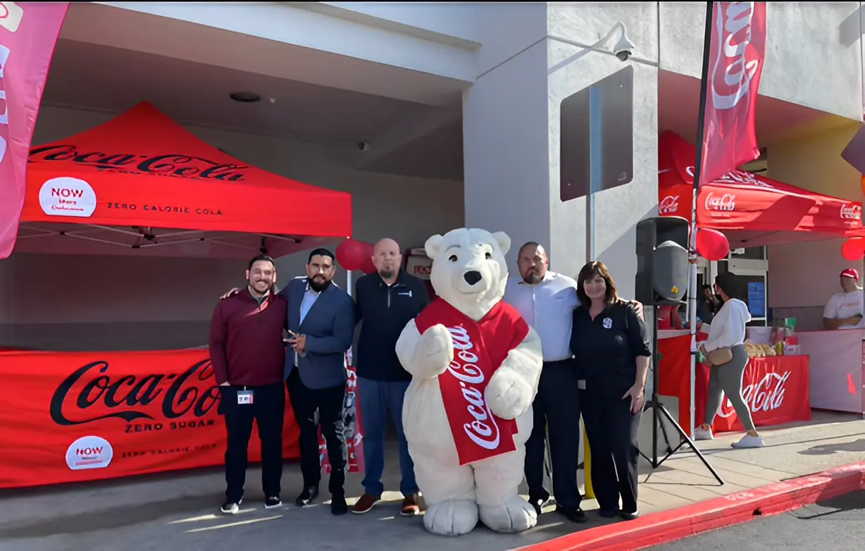 A group of people stands next to a Coca-Cola polar bear mascot in front of a red Coca-Cola tent.