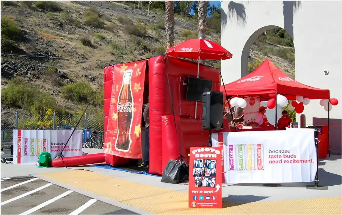 A Coca-Cola themed photo booth with red inflatable walls, banners, balloons, and a display screen. The booth is set up outdoors near a hill.