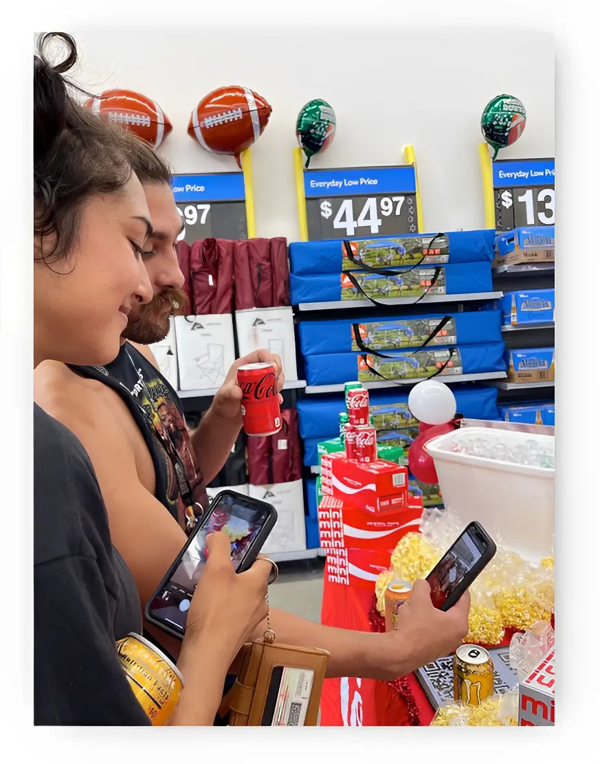 Two people using their phones in a store aisle. There's a display with soda cans, balloons, and a $44.97 price sign in the background.