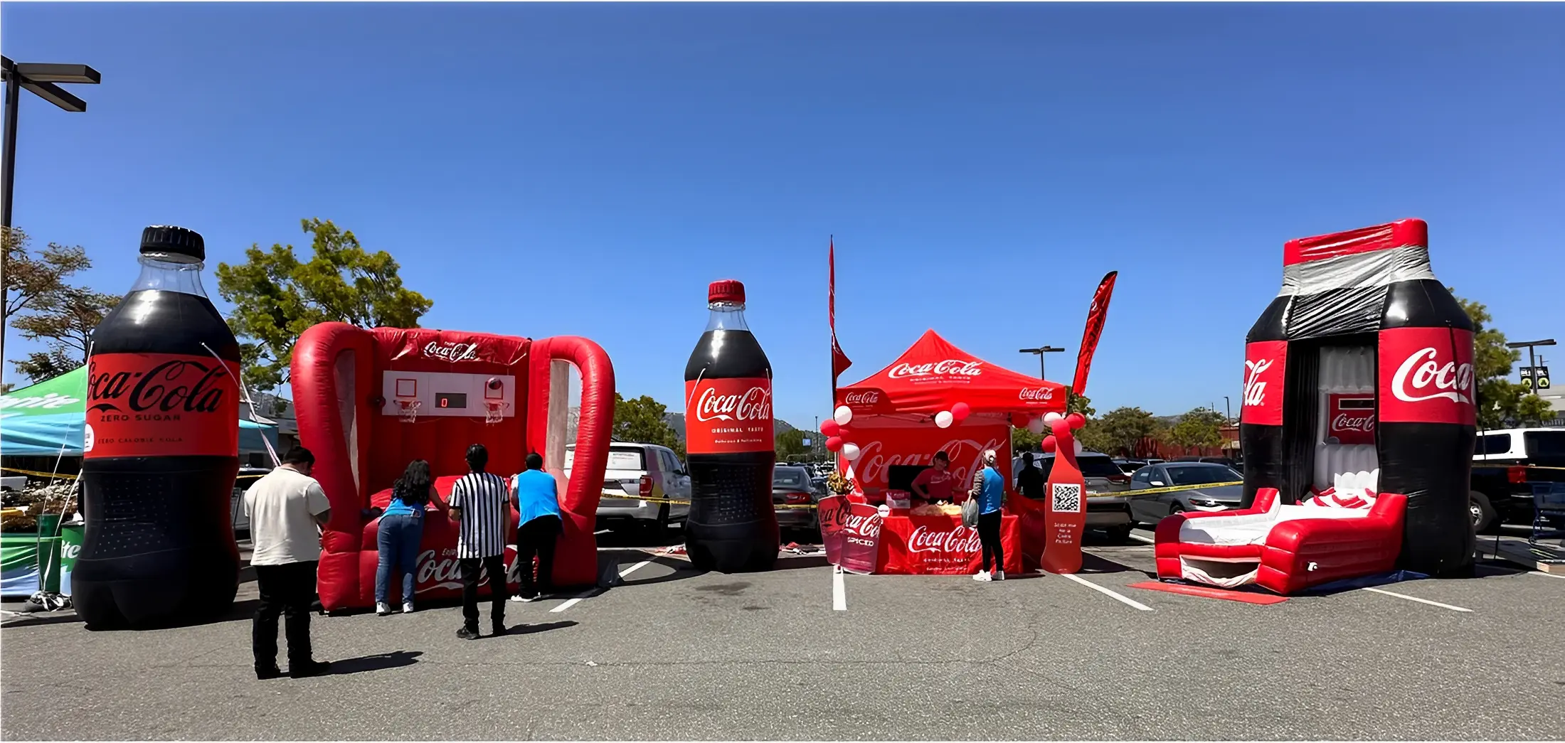 Outdoor display featuring large inflatable Coca-Cola bottles, a basketball hoop, and a branded tent, with people interacting under a clear blue sky.