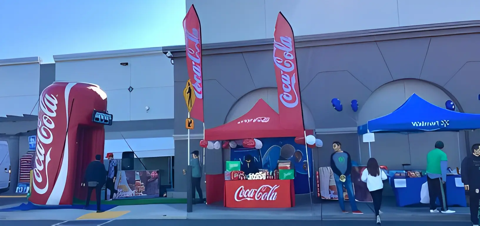 Outdoor Coca-Cola and Walmart event setup with booths and banners. Giant Coca-Cola can display on the left, people gathered nearby.