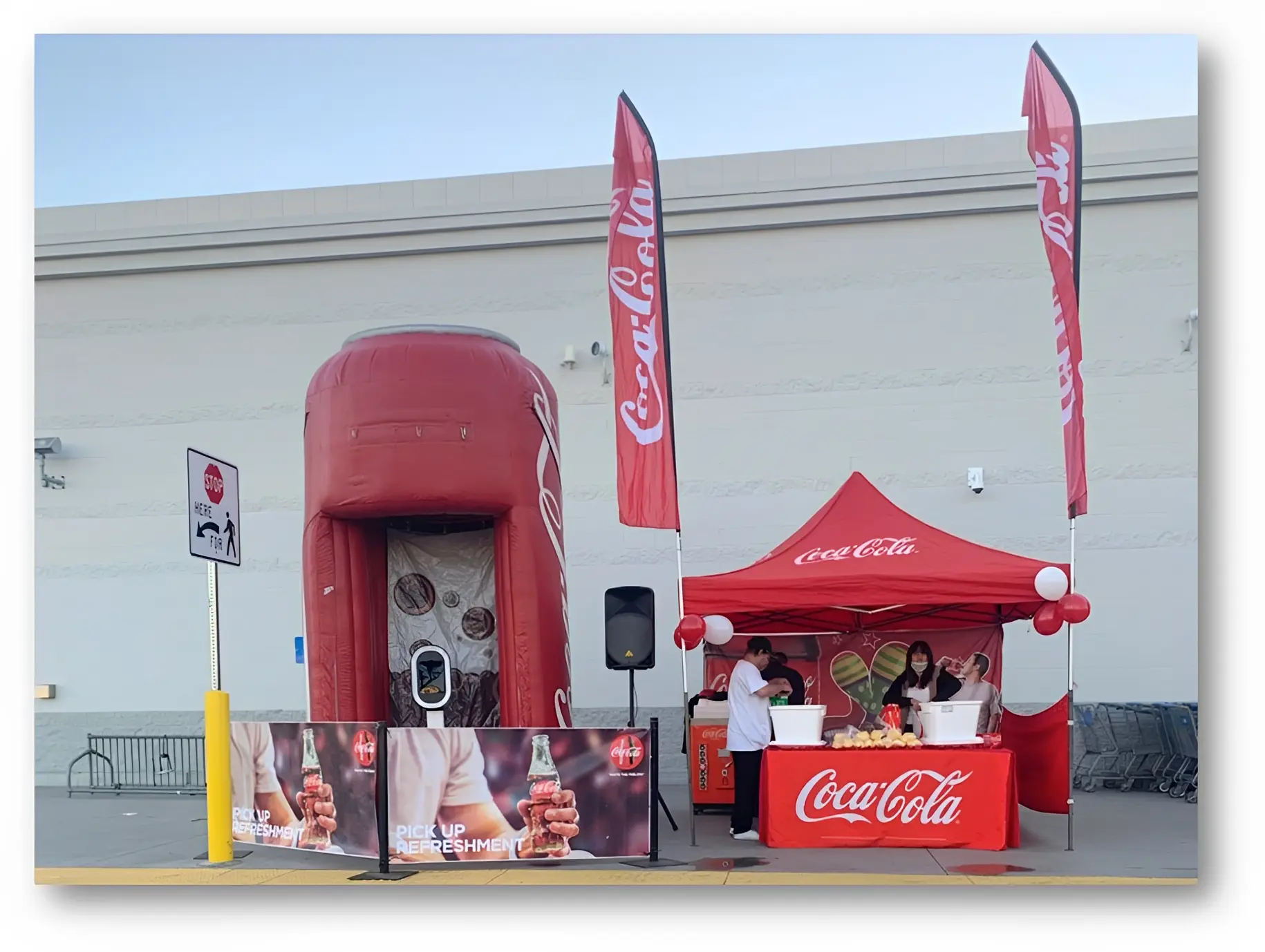 Outdoor Coca-Cola promotional booth with a red tent and inflatable can display. Staff behind a counter with refreshments, red flags, and balloons.