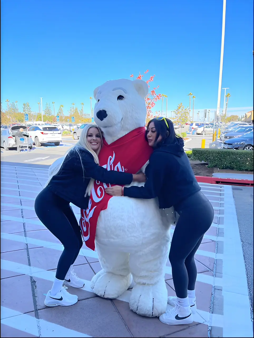 Two women hugging a Coca-Cola polar bear statue outdoors on a sunny day, with cars and trees in the background.