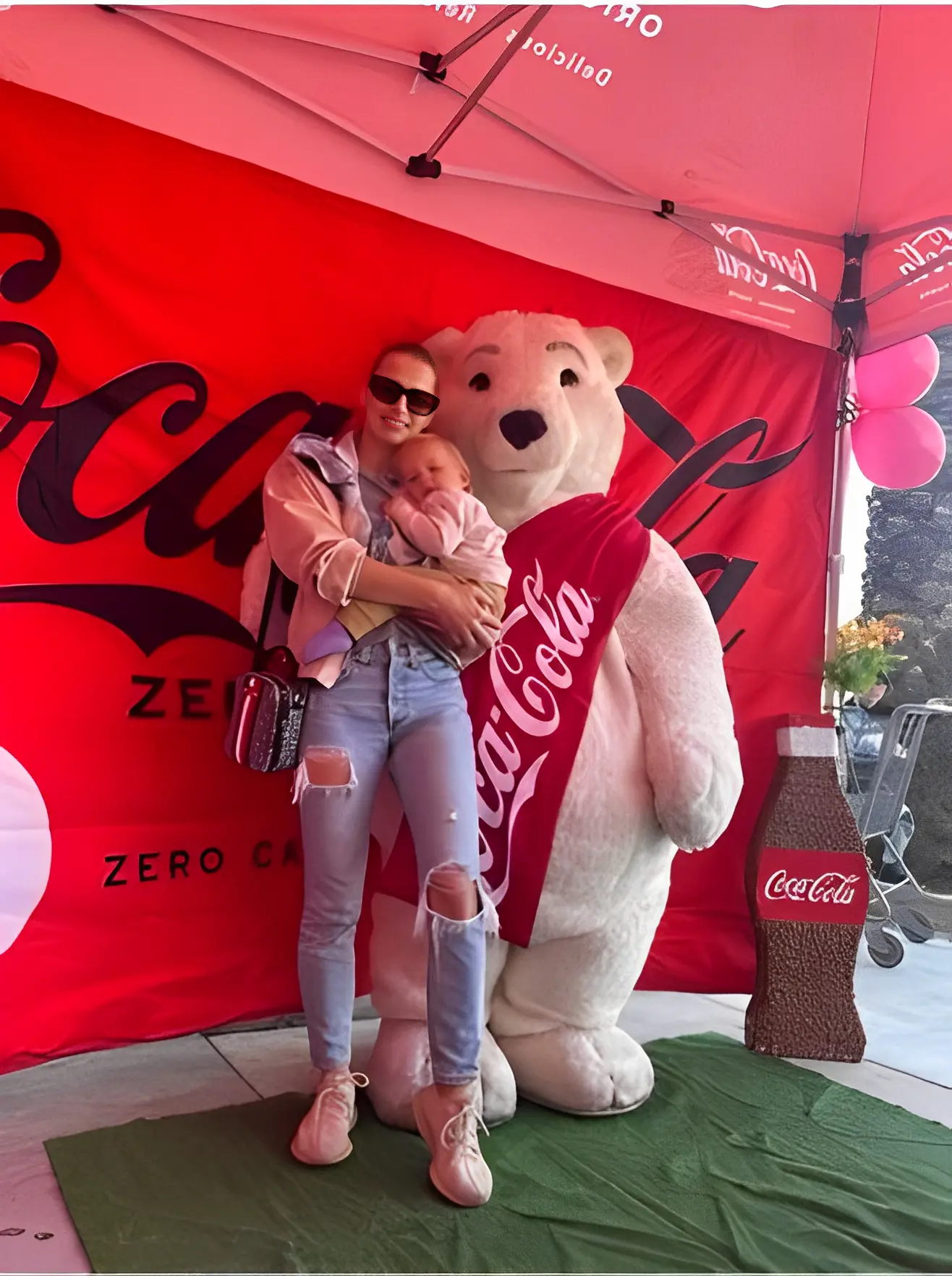 A woman holding a baby stands under a red Coca-Cola tent beside a large Coca-Cola polar bear mascot.