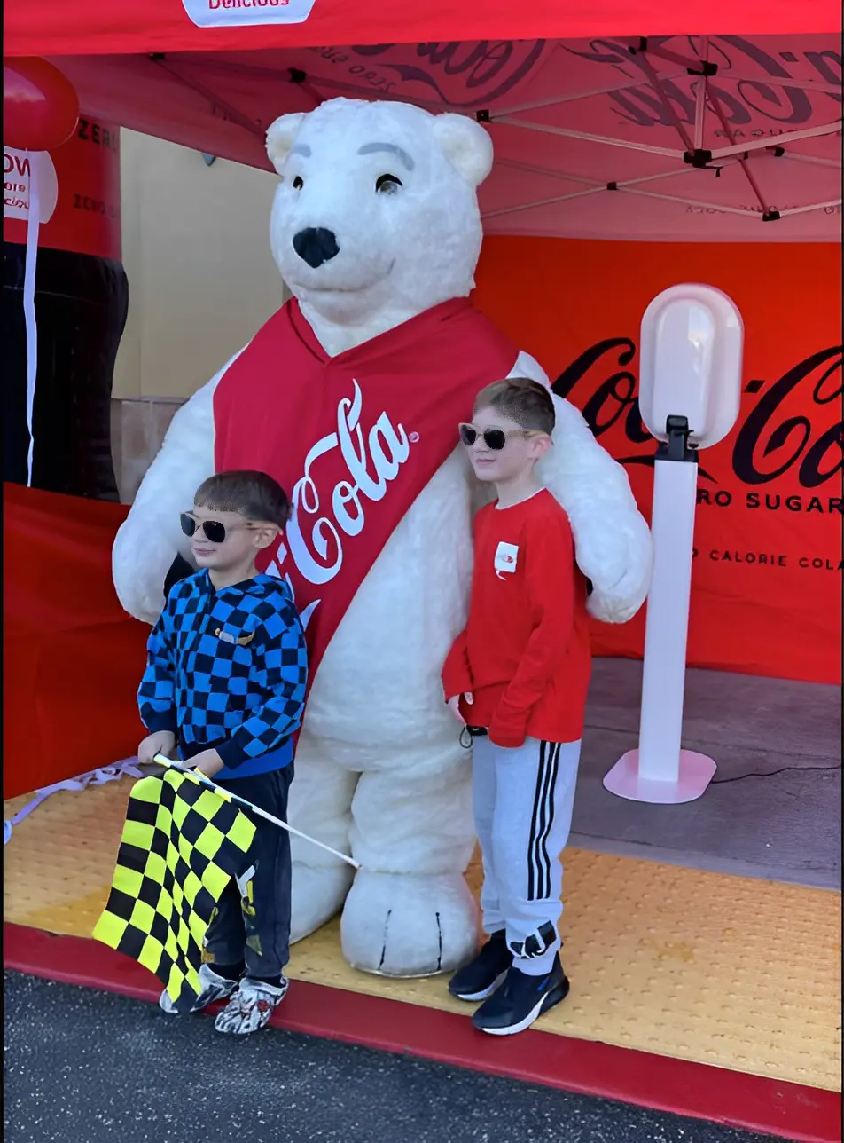 Two children wearing sunglasses pose with a person in a Coca-Cola polar bear costume under a Coca-Cola tent. One child holds a checkered flag.