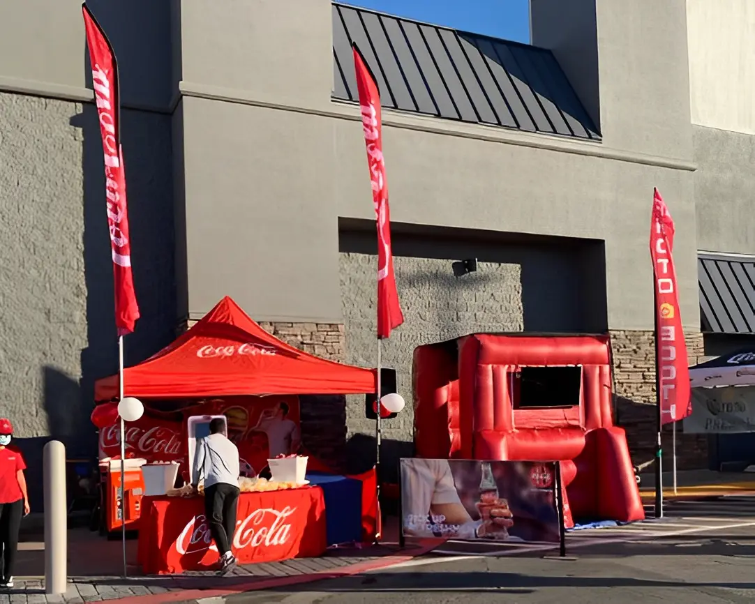 Outdoor Coca-Cola promotional booth with red tent and inflatable display, set up near a building with two tall flags and a banner.