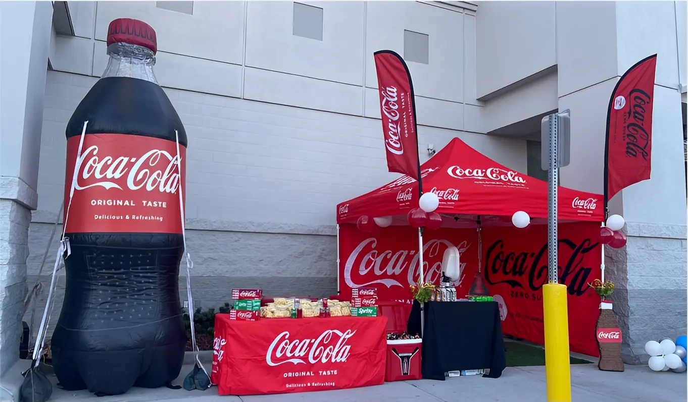 Coca-Cola promotional setup with a large inflatable bottle, branded canopy tent, and tables displaying snacks and drinks outside a building.