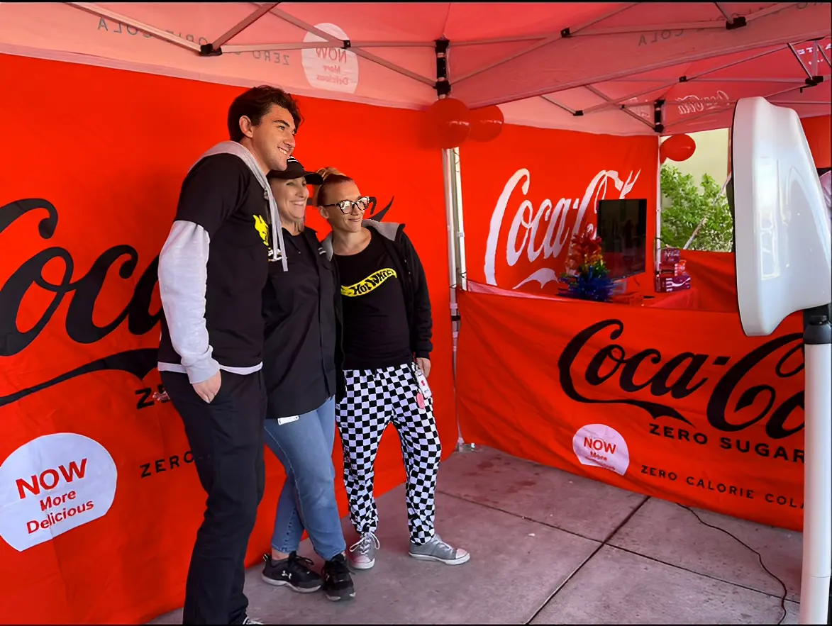 Three people pose for a photo in front of a Coca-Cola branded backdrop under a red canopy.
