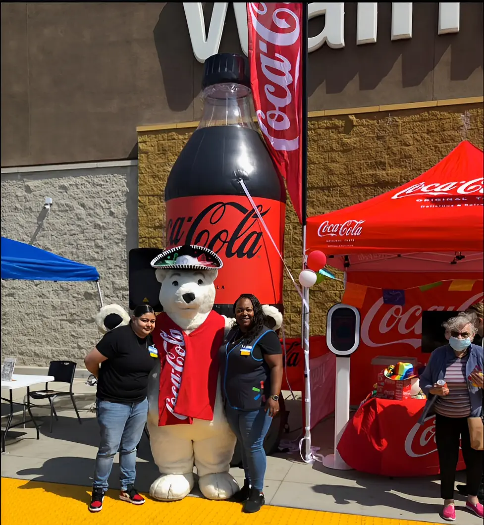 Two people and a person in a polar bear costume stand in front of a large inflatable Coca-Cola bottle and a red canopy.